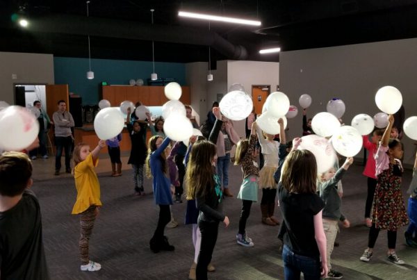 Children holding white balloons indoors, participating in an event.